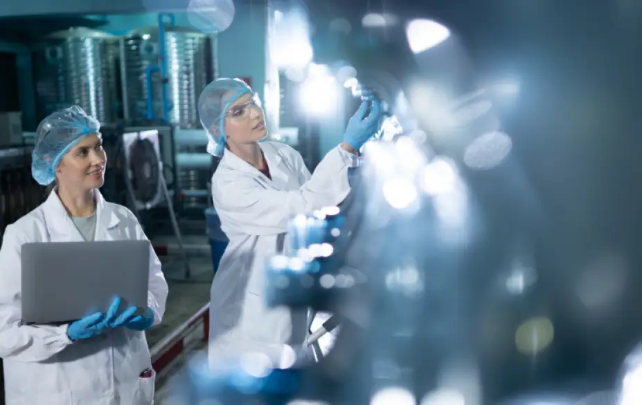 Two female professionals working in a sterile industrial or laboratory environment. Both are wearing white lab coats, blue hairnets, and blue nitrile gloves. The woman on the left is holding an open laptop and looking upward, while the woman on the right is closely inspecting a small bottle or vial that she is holding up to the light. The foreground features a soft, blurred bokeh effect from reflective surfaces, likely glass bottles or containers on a production line. In the background, stainless steel industrial equipment is visible, suggesting a pharmaceutical, food production, or beverage quality control setting. The overall lighting has a cool blue-toned ambiance.